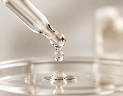 Glass laboratory pipette releasing a drop into a clear dish, on a pale mineral background.